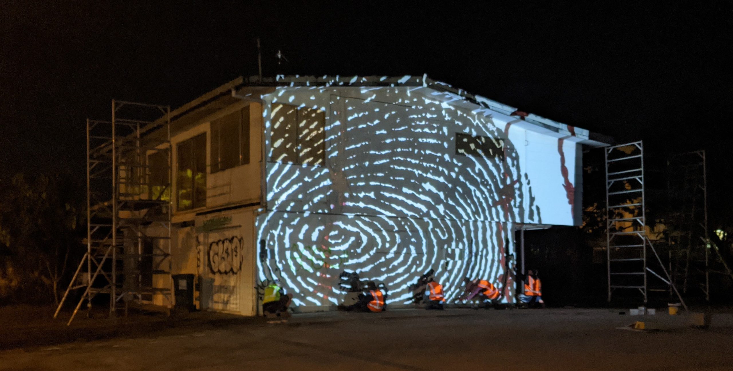 An image of a black thumbprint is projected onto the side of a two storey house. Several pieces of scaffolding are visible. 